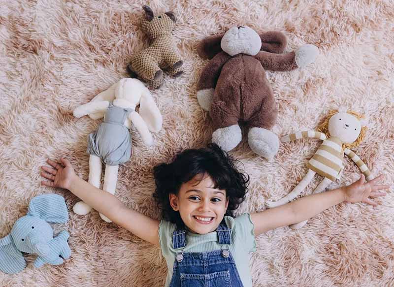 A happy child lies on a soft carpet with arms stretched wide, surrounded by several plush toys in different shapes and styles. The image conveys warmth, comfort, and the emotional appeal of plush toys as cozy companions for children.