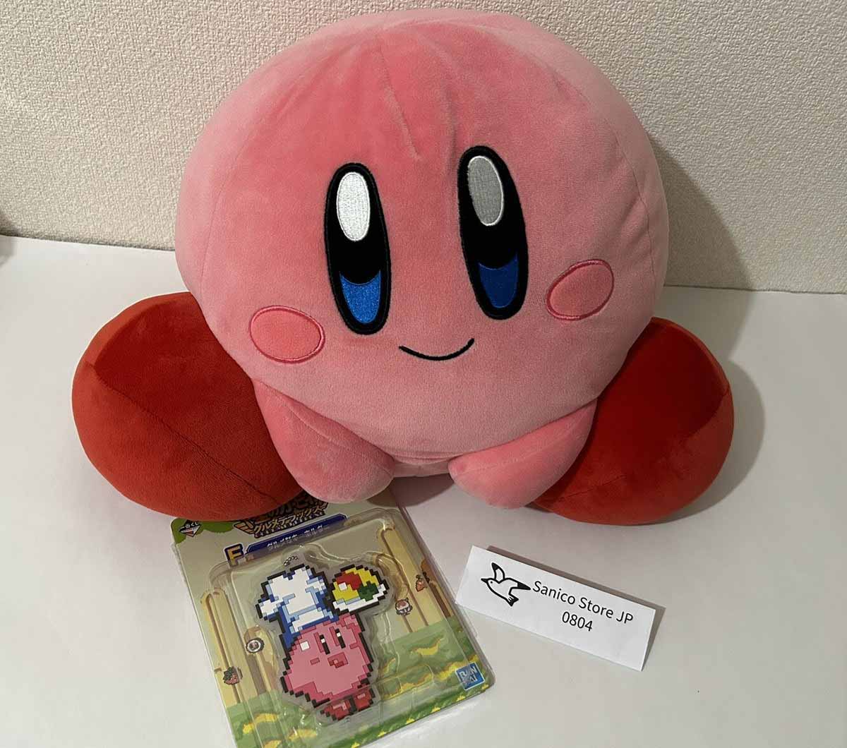 A round pink character plush toy with embroidered facial features and soft red feet, presented alongside retail packaging items for display.