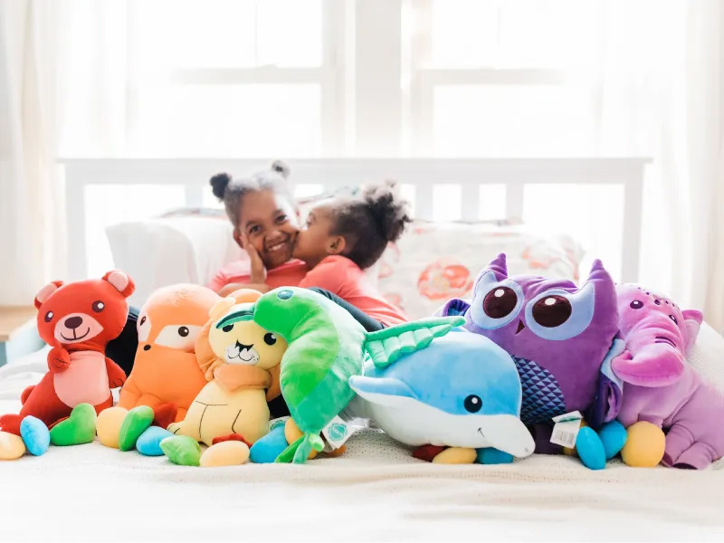 Two children sitting on a bed surrounded by colorful plush toys, showing how stuffed animals provide comfort, fun, and companionship during playtime.