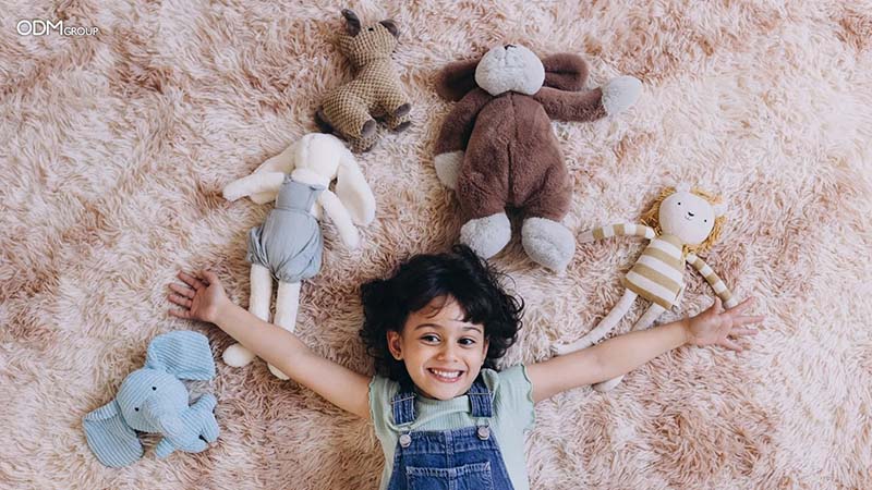 A smiling child lies on a fluffy carpet with arms stretched out, surrounded by several soft plush toys in different animal and doll designs. The image creates a warm, comforting atmosphere and highlights the soothing, child-friendly appeal of plush toys.