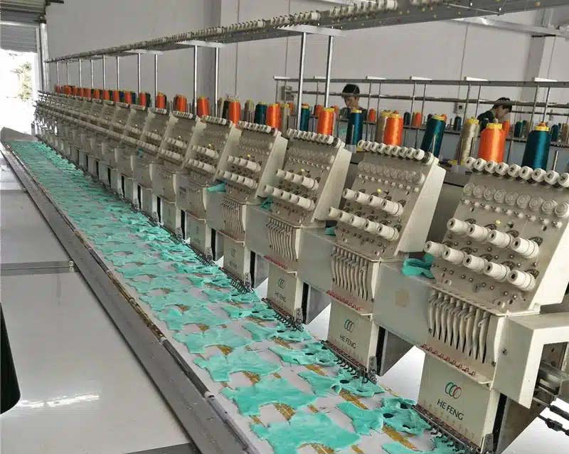 A row of multi-head industrial embroidery machines operating in a factory, used for mass embroidery of plush toy panels before assembly.