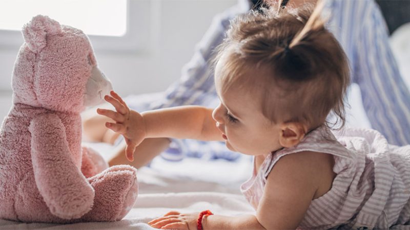 Baby lying on a bed, reaching out to touch a soft pink teddy bear plush toy during playtime.
