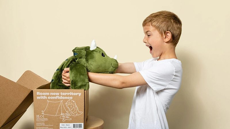 A young boy joyfully holding a green weighted dinosaur plush toy taken from a cardboard box, showing excitement and sensory comfort during play