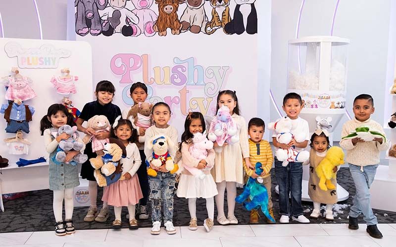 A group of young children standing together at a plush toy store event, each holding a stuffed animal, with colorful plush displays and signage in the background.