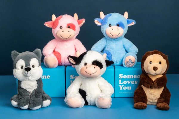 Six plush animal toys arranged in two rows against a blue background, including pink and blue cows, a black-and-white cow, a gray wolf, and a brown monkey, displayed with blue gift boxes that read ‘Someone Loves You.
