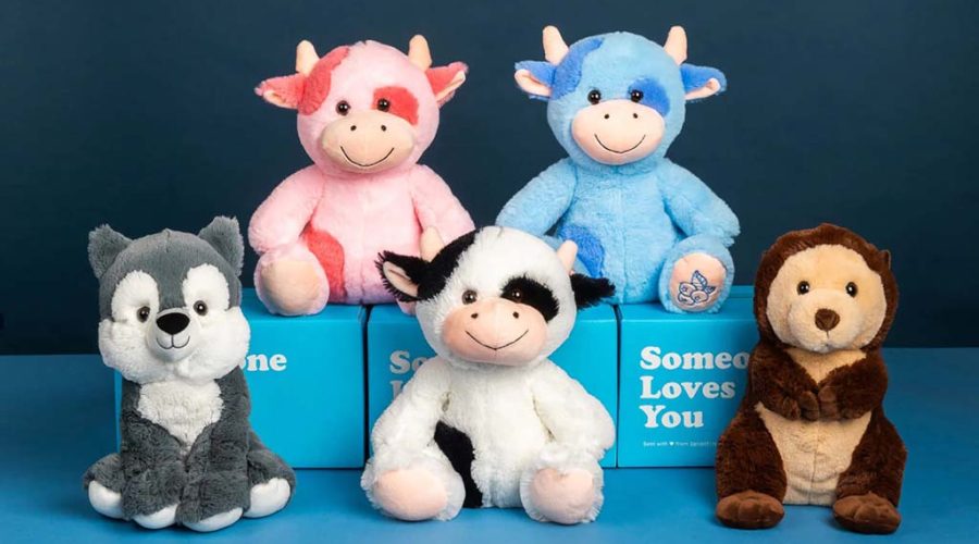 Six plush animal toys arranged in two rows against a blue background, including pink and blue cows, a black-and-white cow, a gray wolf, and a brown monkey, displayed with blue gift boxes that read ‘Someone Loves You.