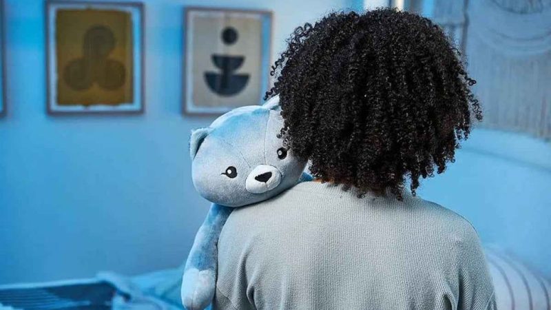 Person holding a blue weighted teddy bear over their shoulder in a softly lit bedroom, conveying comfort and emotional support.