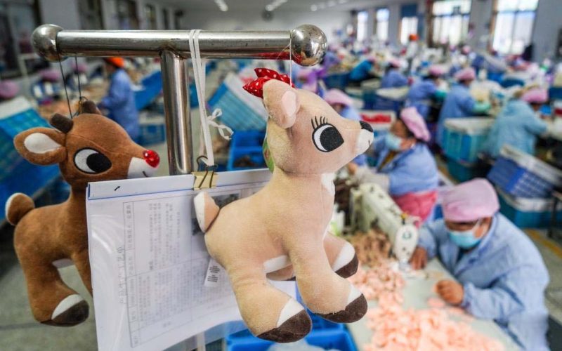 Deer plush toys hanging on a production rack with paperwork, with workers sewing in a busy plush toy factory background.