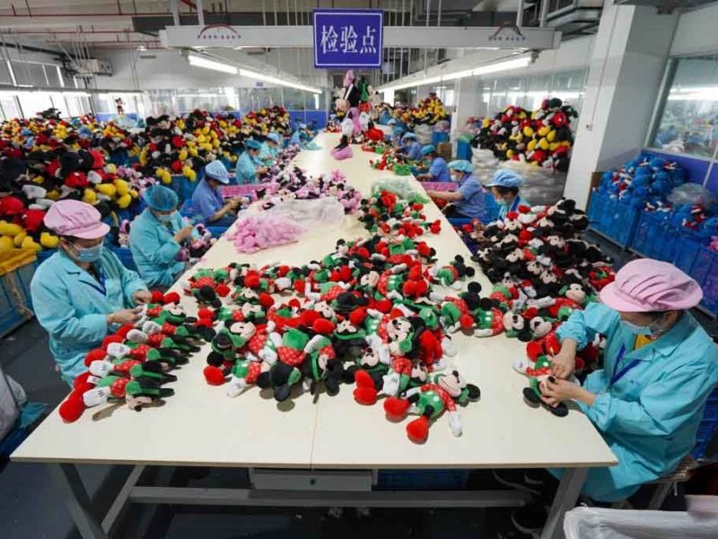 Workers inspecting and sorting stuffed toys at a large factory inspection station with piles of plush animals on tables.