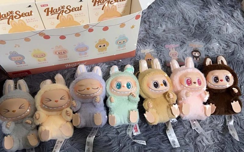Row of pastel-colored big-eyed bunny plush keychain dolls from a blind box series, each with vinyl face details, displayed on a soft gray surface with branded packaging behind.