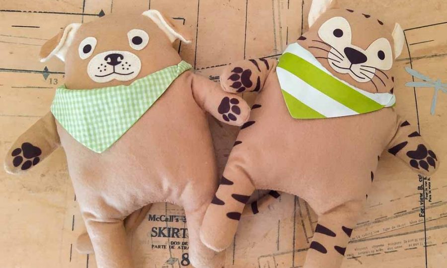 Two handmade plush toys—a dog and a tiger—lying on a sewing pattern background. Both toys have soft tan bodies, printed facial features, paw details, and green bandanas (one gingham, one striped). Their rounded shapes and simple limbs give them a cute, friendly look.
