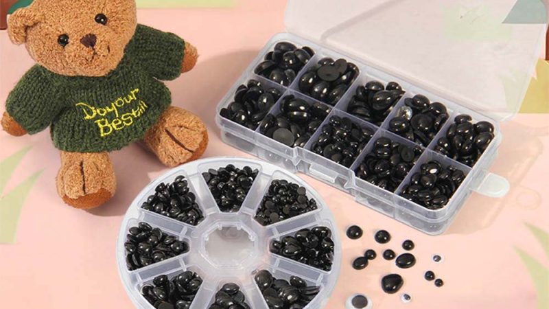 A small teddy bear wearing a green sweater sits beside clear plastic organizers filled with black safety eyes and toy components, used for making or repairing stuffed animals.