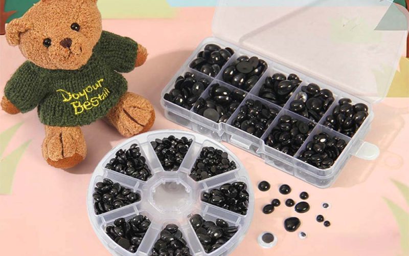 A small teddy bear wearing a green sweater sits beside clear plastic organizers filled with black safety eyes and toy components, used for making or repairing stuffed animals.