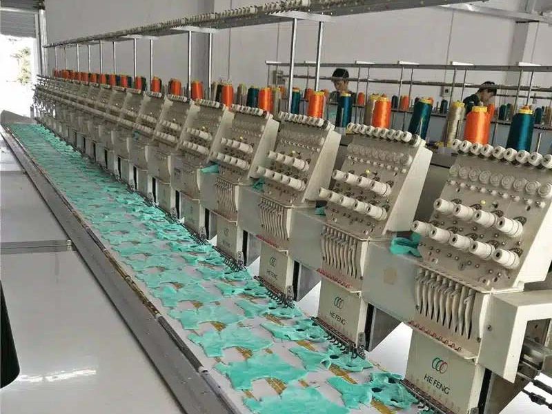 A row of multi-head industrial embroidery machines operating in a factory, used for mass embroidery of plush toy panels before assembly.