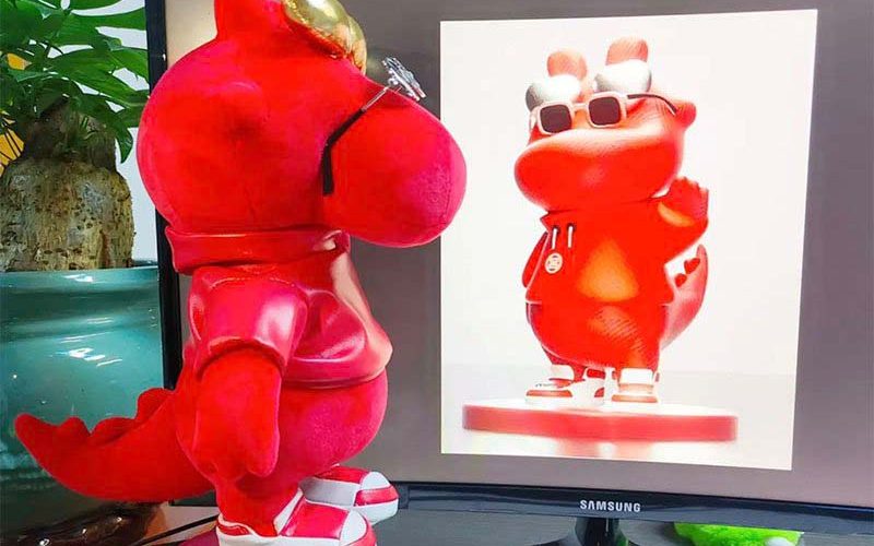 Red dinosaur plush prototype wearing sunglasses and sneakers standing beside a monitor showing the matching character design reference.