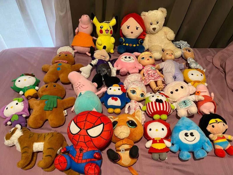 Large collection of mixed plush toys and dolls arranged together on a bed, including animals, cartoon characters, and soft figures