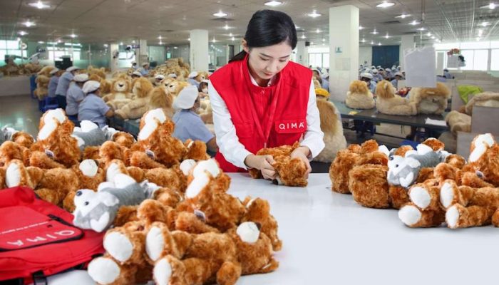 A quality inspector in a red vest examines teddy bears in a plush toy factory, with workers assembling toys in the background.