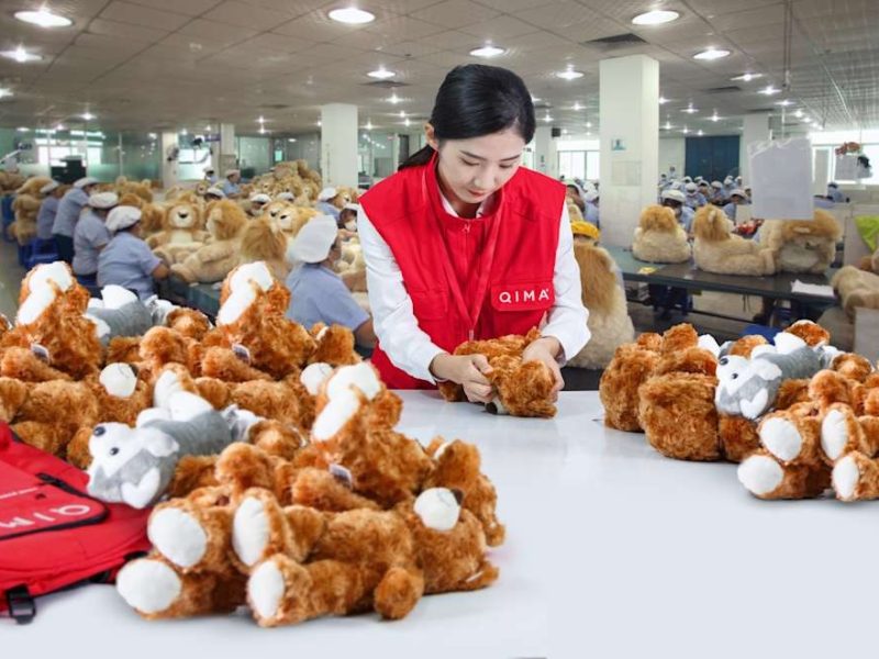 A quality inspector in a red vest examines teddy bears in a plush toy factory, with workers assembling toys in the background.