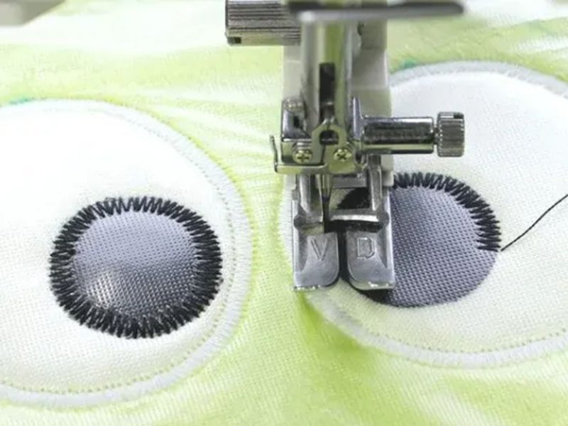 Close-up view of a sewing machine embroidering cartoon eyes onto soft plush fabric, showcasing precision stitching for plush toy detailing.