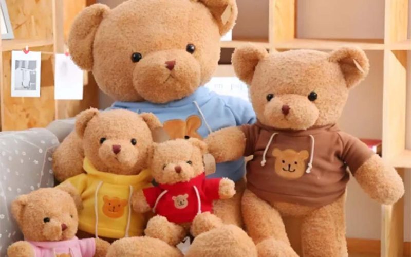 Group of cute teddy bears of different sizes wearing colorful hoodies, sitting together on a cozy couch in a warm indoor setting.