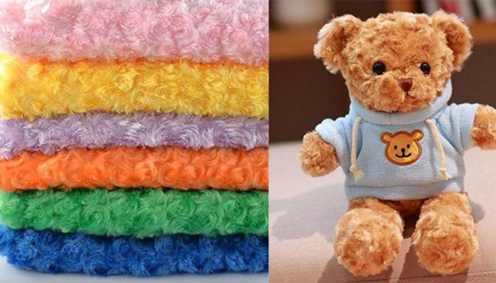 Stack of colorful rose fleece plush fabrics beside a soft teddy bear wearing a blue hoodie, showing material used for custom stuffed toys.