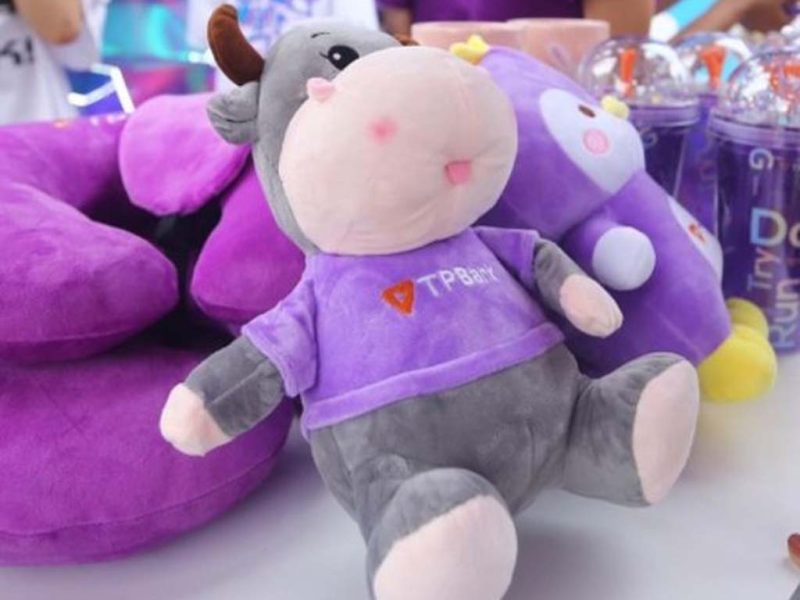 A gray cow plush toy wearing a purple TPBank shirt displayed on a table with other purple promotional items during a marketing event.