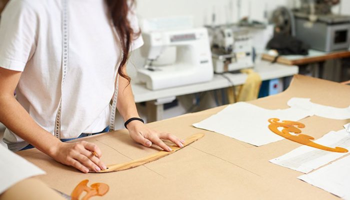 Designer drawing plush toy patterns on brown paper using rulers in a sewing workshop with fabric pieces and sewing machines in the background.