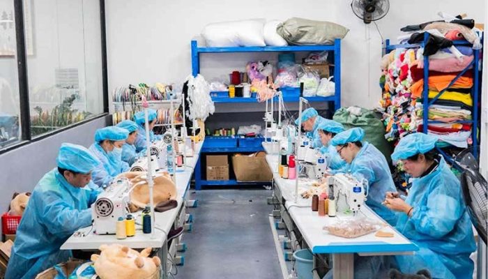 Plush toy factory sewing department with workers in blue protective uniforms operating industrial sewing machines, surrounded by fabric shelves, stuffing materials, and plush components during stuffed animal production.