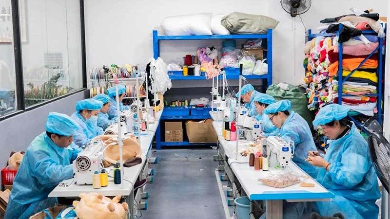 Plush toy factory sewing department with workers in blue protective uniforms operating industrial sewing machines, surrounded by fabric shelves, stuffing materials, and plush components during stuffed animal production.