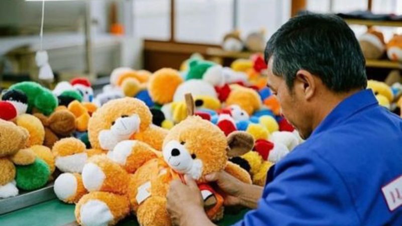 Factory worker inspecting orange teddy bears among colorful plush toys during quality control at a plush manufacturing facility.