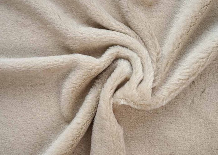 Beige short-pile plush fabric with smooth velvety texture, shown twisted to highlight its softness and dense fiber surface.