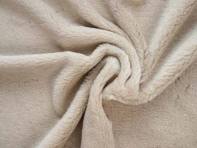 Beige short-pile plush fabric with smooth velvety texture, shown twisted to highlight its softness and dense fiber surface.