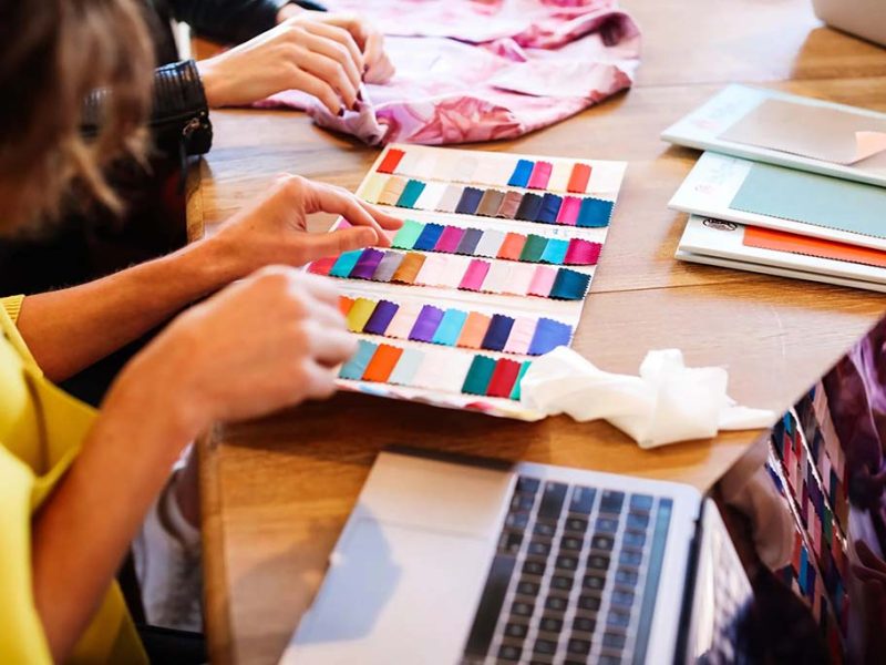 Two people reviewing a fabric color swatch book at a table, comparing textile samples for product design, with a laptop and notebooks nearby during a material selection discussion.