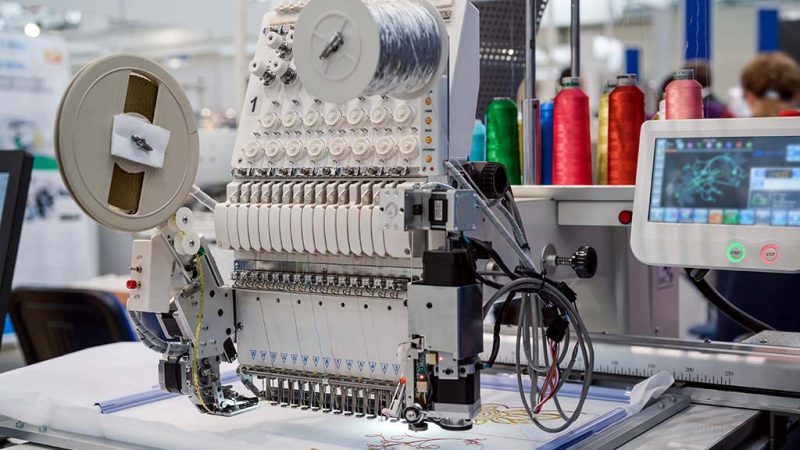 A modern computerized embroidery machine with multiple needles and colorful thread spools stitching a detailed design onto fabric.