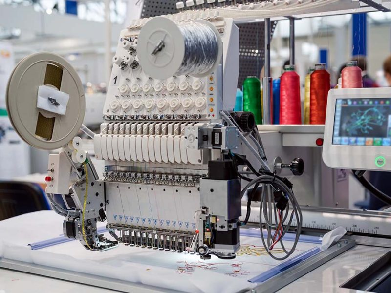 A modern computerized embroidery machine with multiple needles and colorful thread spools stitching a detailed design onto fabric.