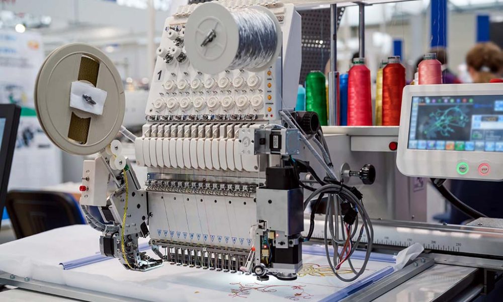 A modern computerized embroidery machine with multiple needles and colorful thread spools stitching a detailed design onto fabric.