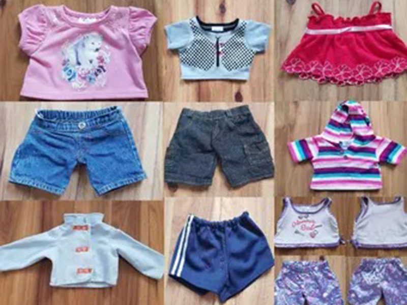 A collection of assorted doll and stuffed animal clothes laid out on a wooden floor, including shirts, shorts, hoodies, dresses, and patterned outfits in various colors and styles.