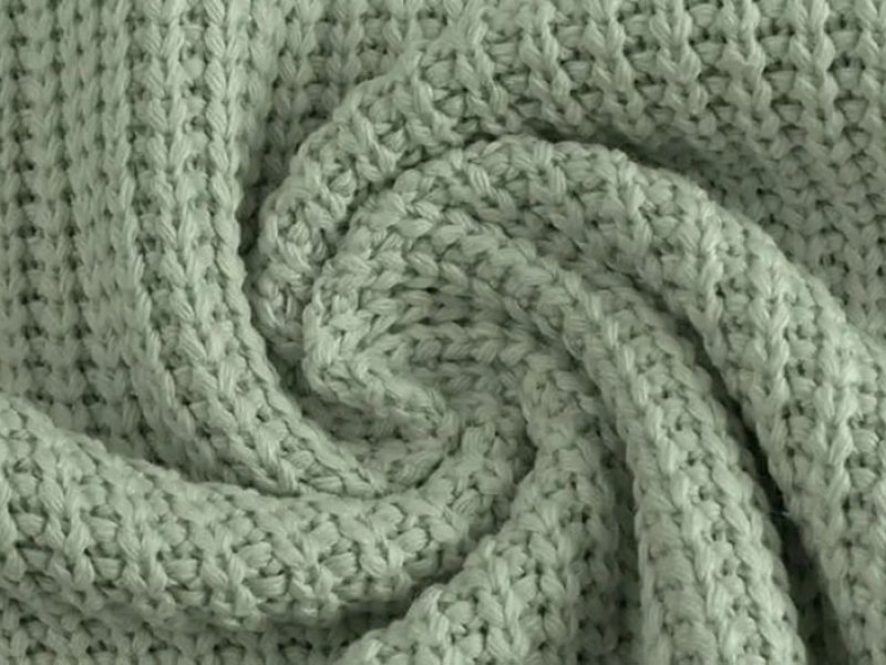 Close-up of soft sage green knitted fabric with a waffle-like weave pattern, often used for plush toy clothing or textured textile designs.