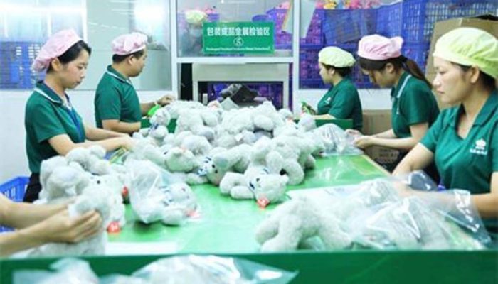 Factory workers in green uniforms and caps package soft plush toys into plastic bags on a green assembly table.