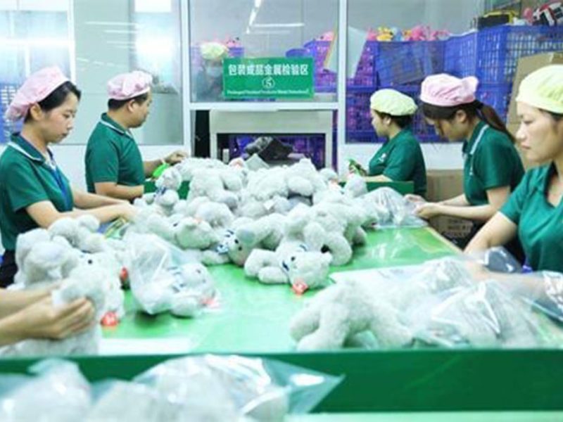 Factory workers in green uniforms and caps package soft plush toys into plastic bags on a green assembly table.