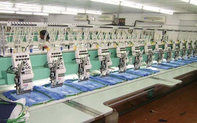 A row of automated multi-head embroidery machines simultaneously stitching designs onto multiple fabric pieces in a factory.