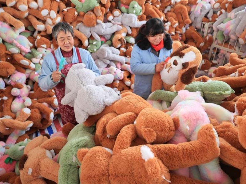 Two workers inspecting and grooming large plush toys in a factory filled with colorful stuffed animals ready for packaging.