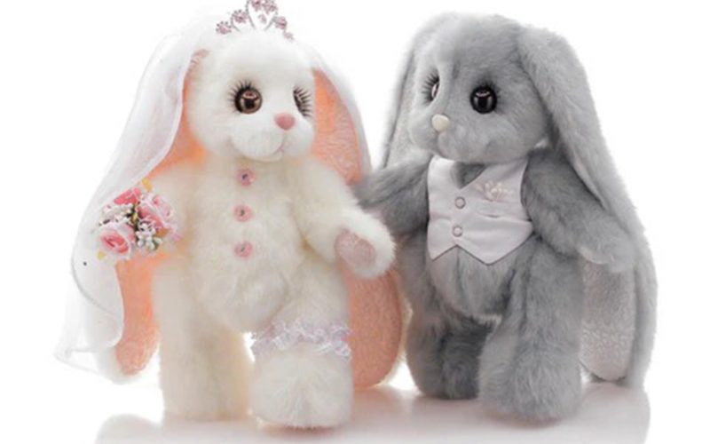 Two plush bunny toys dressed as a bride and groom, with the white bunny wearing a veil and holding a bouquet, and the gray bunny in a vest suit.