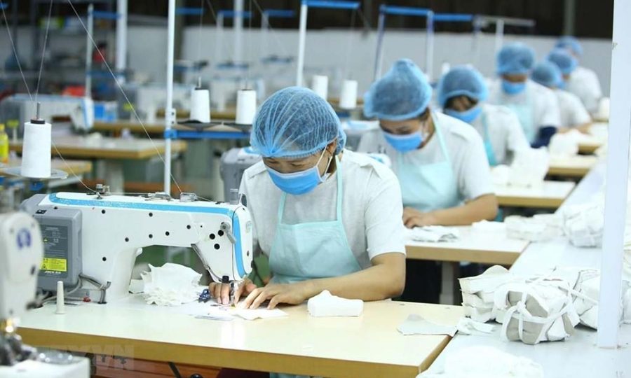 Factory sewing operators wearing masks and hair nets working at industrial sewing machines on a production line, stitching fabric pieces in a clean manufacturing environment.