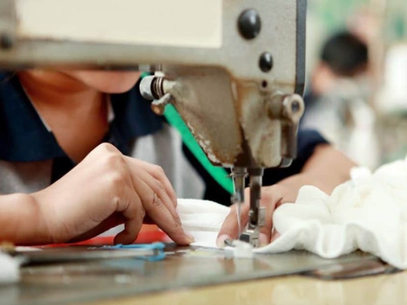 Close-up of a worker using an industrial sewing machine to stitch white fabric, showing hands guiding the material during plush toy production.