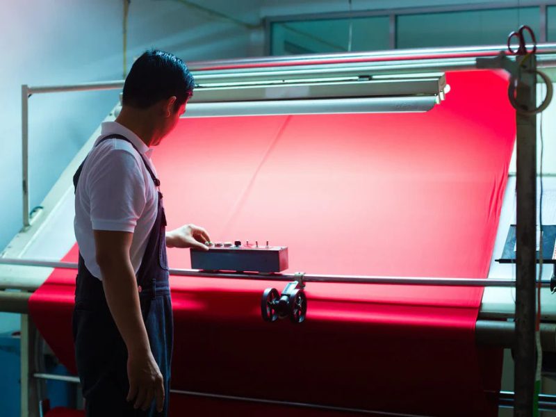 Technician inspecting bright red fabric on a textile production line under controlled lighting to ensure quality and consistency.