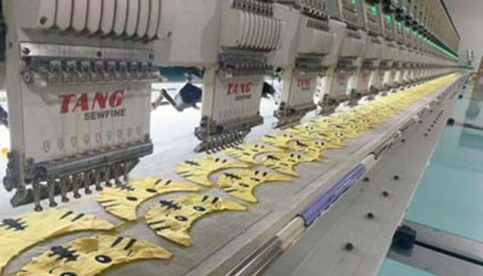 Industrial embroidery machines stitching yellow plush fabric pieces with cartoon faces in a plush toy factory production line.