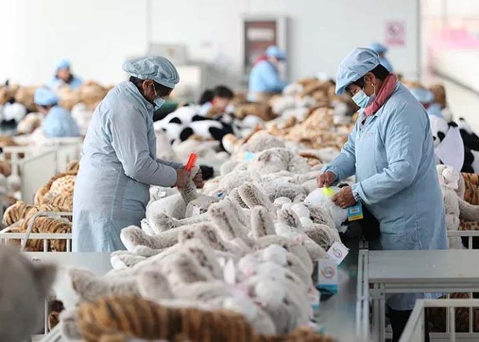 Workers in protective uniforms inspecting and grooming plush toys inside a large stuffed-animal manufacturing factory.